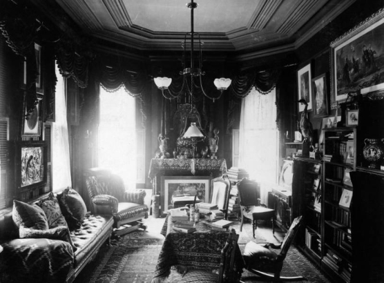 Interior view of the F. J. Bancroft house in Denver, Colorado; decor includes Oriental rugs, a hanging lamp, a taxidermied heron, ewers, a Buddha, framed art, and upholstered chairs.