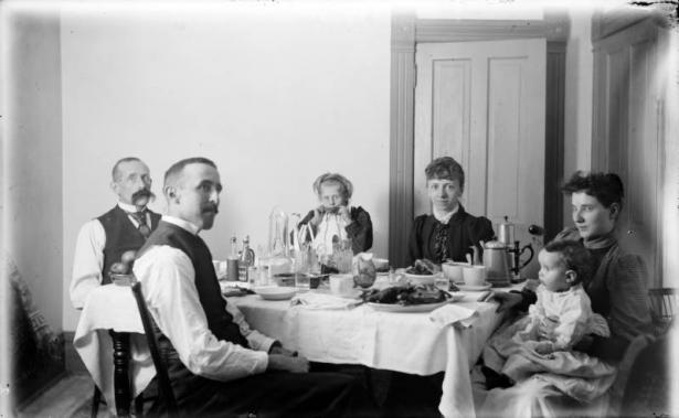 Two men, two women, a girl, and a baby are seated for a meal, probably in Denver, Colorado. Items on the table include: a tablecloth, plates, cups, a coffee pot, a water pitcher, glasses, silverware, and food.