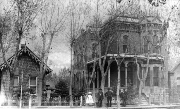 View of residences in the Auraria neighborhood of Denver, Colorado. One has a covered porch, mansard roof, a widow's walk, and bargeboard. Men and women pose.