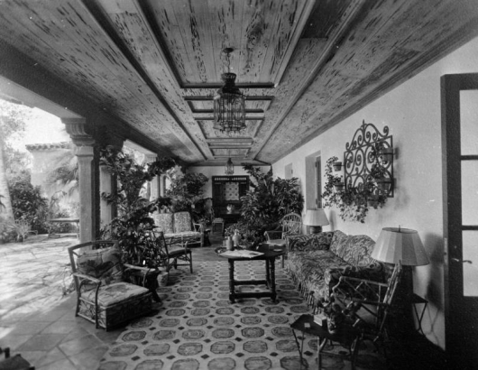 View of a covered patio in Denver, Colorado. Coffered ceilings, wrought iron lamps, couches, chairs, potted plants, and a tile floor comprise the decor. An open plaza is to the side.