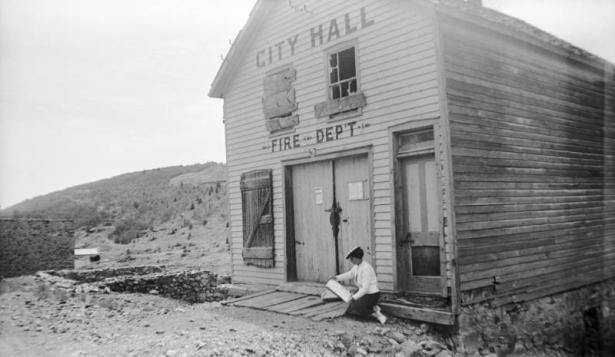 A woman (Muriel Wolle?) is sitting at the entryway of the City Hall and Fire Department building in Nevadaville, Colorado. The name of the building is painted on the facade, and surrounds two broken and boarded up window bays. The woman is holding a sketchpad in her hands.