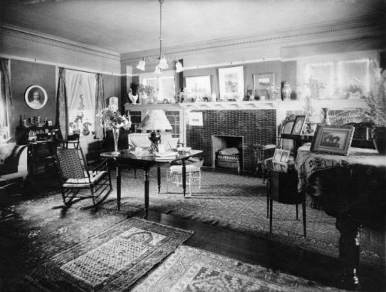 Interior view of a house in Denver, Colorado; decor includes Oriental rugs, rocking chairs, a fireplace, lamps, flower vases, porcelain, photos, and a piano.