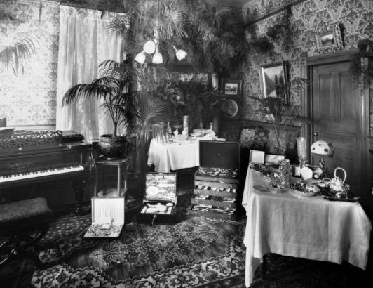 Interior view of a home in Denver, Colorado; decor includes an Oriental rug, console grand piano, potted plants, cases of silver dinnerware, a toilet set, and dishes.