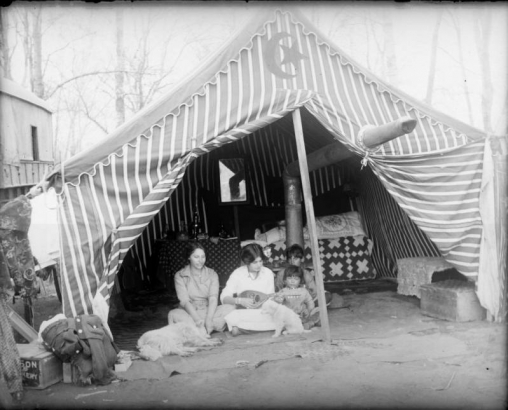 Portrait of women, girls, and dogs in a tent in (probably) Denver, Colorado; one plays a mandolin. A star / crescent motif is on the striped canvas; embroidery and quilts are inside.