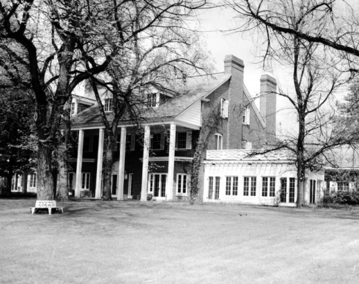View of a house owned by Temple Hoyne Buell probably in Denver, Colorado. The two-story brick house has a pitched roof, dormers, chimneys, columns, and covered entryway. A one-story sun room is nearby.