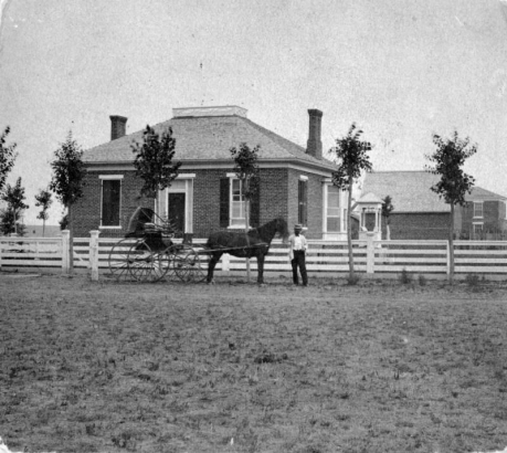 Cyrus H. McLaughlin poses by a horse-drawn buggy near the Alida Burton house at 100 West Colfax Avenue in the Civic Center neighborhood of Denver, Colorado. The one-story brick house has a pitched roof, chimneys, and a balustrade.