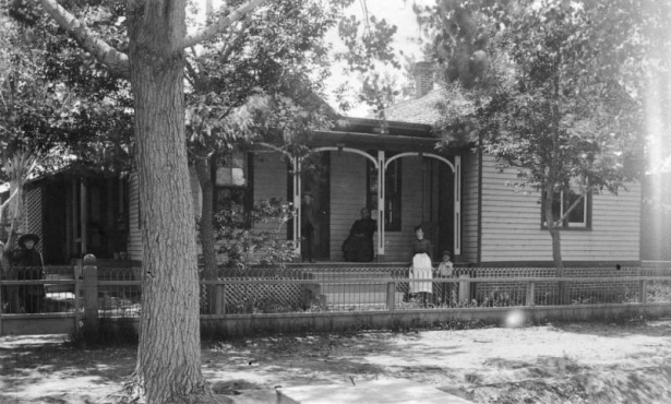 Boys, women, and a girl pose outdoors near the John L. Dailey house at 4th (Fourth) Avenue and Broadway in Denver, Colorado. The wood frame house has a covered porch and is surrounded by a metal fence.