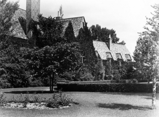 View of the George W. Gano house in Englewood (Arapahoe County), Colorado. The two-story brick house has a hip roof, chimney, and dormers. Trees and bushes are nearby.