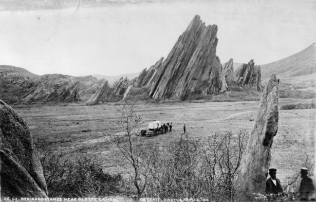 View of a mule drawn wagon with supplies set behind it, possibly the photographer's, near large jagged sandstone rock formations in what is now Roxborough State Park, Douglas County, Colorado. Shows a man by the wagon; two men pose by a small rock formation in the foreground.