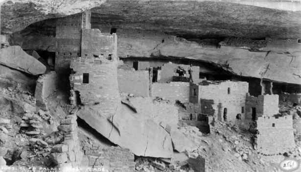 A man poses in Cliff Palace, a Native American (Anasazi) cliff dwelling at Mesa Verde National Park, in Montezuma County, Colorado. Shows masonry ruins and a stone overhang.