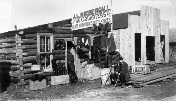 Two men and a dog are outside a log store for miners in Fremont (Cripple Creek, Teller County), Colorado. The sign above the store reads "J.L. Niebergall, Headquarters for Gents Furnishings Boots & Shoes."  Shirts, pants, jackets, gloves, socks, shoes and boots are arranged on boxes and hung on beams outside the cabin.  A sign outside the cabin includes price list. Suspenders hang in the window. A sign: "Lodging," is on a stack of building materials beside a plank building under construction.