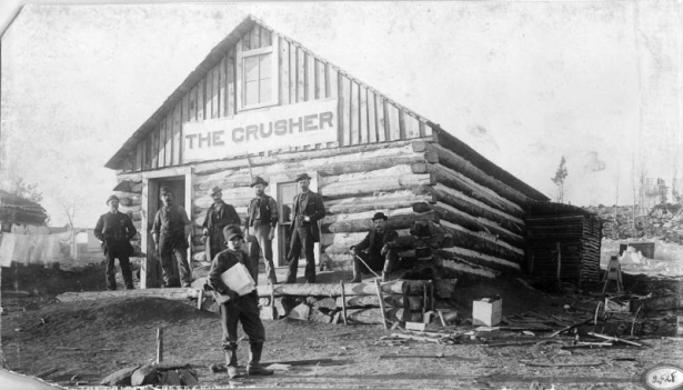 Six men are gathered on the front deck of the The Crusher, the first newspaper in Fremont (Cripple Creek, Teller County), Colorado.  Oakley Spell, a young boy, holds a bundle of newspapers under his arm; a sign on the log building reads: "The Crusher."