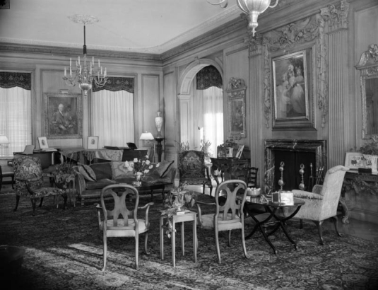 Interior view of the living room in the Lawrence Cowle Phipps house at 3400 Belcaro Drive in the Belcaro neighborhood of Denver, Colorado. Chairs, tables, a sofa, fireplace, a chandelier, and portraits comprise decor.