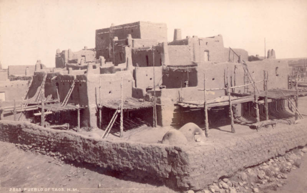 View of Native American (Taos) Taos Pueblo, New Mexico; shows adobe buildings with wood decks, adobe wall and ovens.