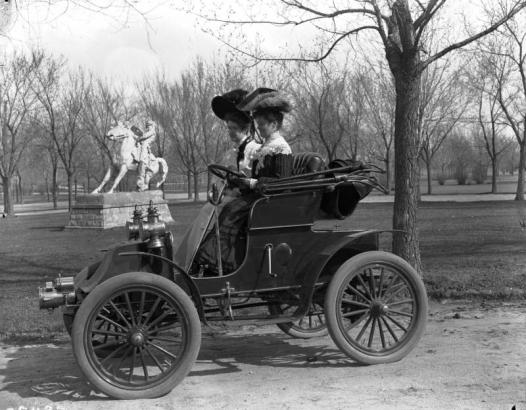Women ride in an early automobile in City Park, Denver, Colorado. They wear dresses and hats decorated with feathers. A statue, 'The Cowboy,' created by Alexander Phimister (A. P.) Proctor, is in the distance.