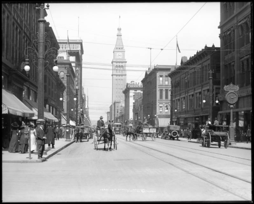 View northwest from California Street down 16th (Sixteenth) Street, Denver, Colorado; shows automobiles, horse-drawn wagons, delivery wagons & carriages, street railway car & tracks, Daniels & Fisher tower, streetlights with electric tramway cables, Stark street clock, pedestrians on sidewalk, Gano-Downs (721-739 16th & corner of Stout), A.T. Lewis & Son (800-818 16th & corner of Stout), May Company building.
