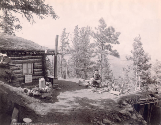 A prospector and guide, with a pipe and a dog and puppies, sits by his log cabin in Teller County, Colorado. The cabin is built into a hill and has a sod roof. A cast-iron wood stove is outside the cabin, a sign reads "Burros for hire." Shows a sawhorse and a can with a spout.