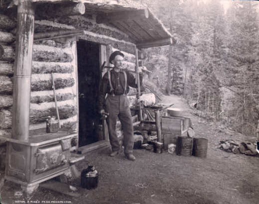 A prospector with a sledgehammer stands outside his chink and log cabin in Teller County, Colorado; shows a cast-iron stove outside the door of the cabin, a jug, broom, lunch pail, pans, cans, and a dog and her puppies under a table.