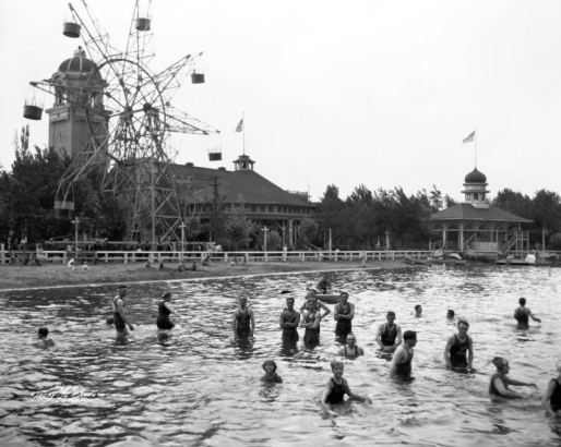 Men, women and children swimmers wade in Lake Rhoda at Lakeside Amusement Park in Lakeside (Jefferson County), Colorado. A lifeguard sits in a row boat and several people lounge on the beach. The Staride ferris wheel, a pavilion and the Casino Tower entrance are in the background.