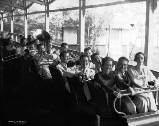 Tom Gerun and members of his orchestra are seated in a roller coaster car at Lakeside Amusement Park in Lakeside (Jefferson County), Colorado. Tom Gerun and two women sit in the front of the car. The musicians are crowed in the rear seats and have their instruments in hand.