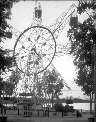 A view of the Staride ferris wheel at Lakeside Amusement Park in Lakeside (Jefferson County), Colorado. A man sits in a ticket booth at the base of the ride. Lake Rhoda is in the distance.