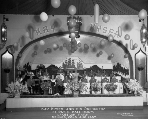 Band leader Kay Kyser stands on stage with his thirteen piece orchestra and two singers at the El Patio ballroom, Lakeside Amusement Park, Lakeside (Jefferson County), Colorado. Across the top of the stage letters read: "Kay Kyser," and balloons are strung from the ceiling. The stage backdrop is decorated with paintings of trees, islands and tall ships. The front of the stage is decorated with baskets of flowers.