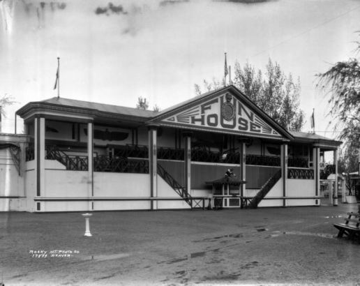 Exterior view of the Fun House at Lakeside Amusement Park, Lakeside (Jefferson County), Colorado. The building has French colonial features including paired exterior staircases, a pavilion roof and thin wooden colonettes. Art Deco designs decorate the columns and the sign reading "Fun House." A face mask and smiling clock are located on the gable. A clown head is attached to the porch, and a monkey figure sits on top of the ticket booth.