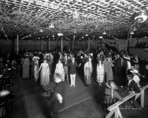 Dancers crowd the floor of the El Patio Ballroom at Lakeside Amusement Park in Lakeside (Jefferson County), Colorado. A man and four women in satin dresses are lined up in one corner of the room while couples occupy the rest of the floor and the tables surrounding dance area. The hall is decorated with an ivy covered trellis and tulip light fixtures.