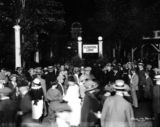 Nighttime view of men and women who crowd around the edge of a sidewalk referred to as "Flapper Lane" at Lakeside Amusement Park in Lakeside (Jefferson County), Colorado. Most of the women have short bobbed haircuts, and the men wear caps and straw hats. Two girls walk arm-in-arm, one wears knickers. A policeman stands next to a column. A tower in the distance is dimly illuminated by lightbulbs.