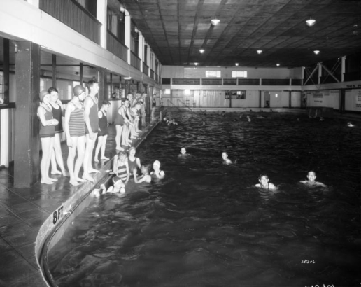Boys and girls stand at the edge of and swim in the swimming pool at Lakeside Amusement Park in Lakeside (Jefferson County), Colorado. The boys and girls both have full bathing suits, and many of the girls wear bathing caps.