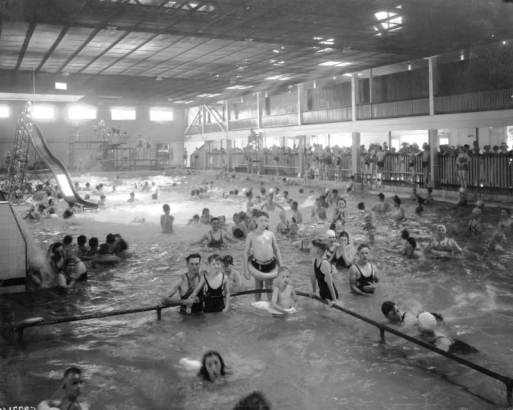 Men, women and children swim in the indoor pool at Lakeside Amusement Park in Lakeside (Jefferson County), Colorado. A girl prepares to go down a slide, and boys and men line up on a platform to use the diving board. Spectators lean against a fence.