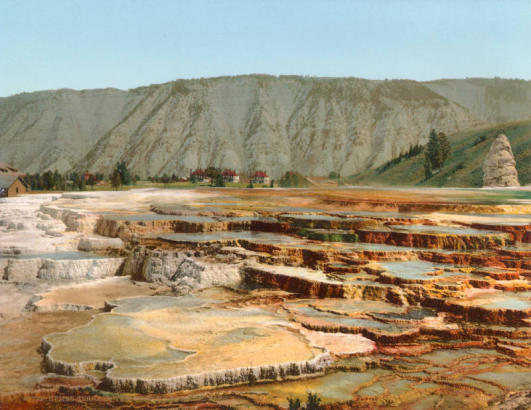 View of Hymen Terrace at Mammoth Hot Springs, Yellowstone National Park, Wyoming. Shows thermal pools and terraces, the Liberty Cap, an extinct hot spring cone, and two-story buildings with red roofs, probably Fort Yellowstone.