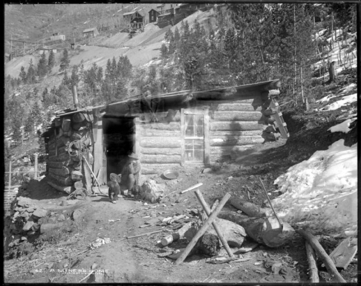 Shows a miner sitting in doorway of his log cabin (lean-to) built into mountainside with glass window frame; includes two dogs, various tools around entrance, wood chopping area, and other mine buildings and structures in background.