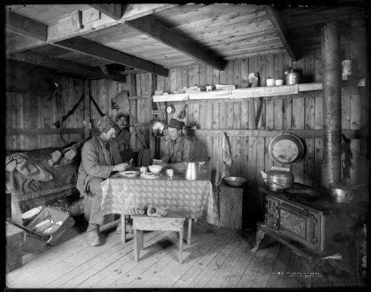 Interior view of a miner's home; includes two men playing cards at table and a miner stretched out on cot; shows wooden framework of house, shelving, wood stove, cooking utensils and gold pan.