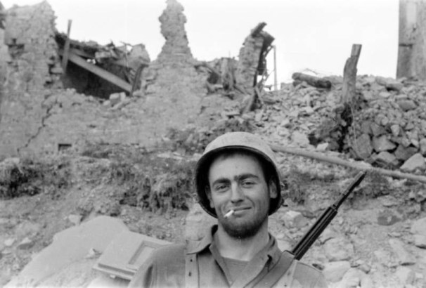 John W. Haines, III, of Company I, 87th Mountain Infantry Regiment, Tenth Mountain Division, faces the camera and smiles with a cigarette in his mouth and a rifle on his back.  Behind him are the ruins and rubble of Pietra Colora. Haines was later killed in action.