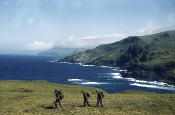 A trio of soldiers from the 87th Mountain Infantry Regiment on patrol loaded with full rucksacks and rifles crosses the muskeg. The sea is beyond them. View is toward the north from near Cape St. Stephen.