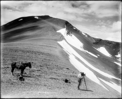 View of summit at Treasure Mountain, Gunnison County, Colorado, with L.C. McClure behind his camera & tripod; horse stands beside unpacked saddle bags; additional camera case, equipment, and probably crate of glass plates are near McClure; clouds top the peaks.