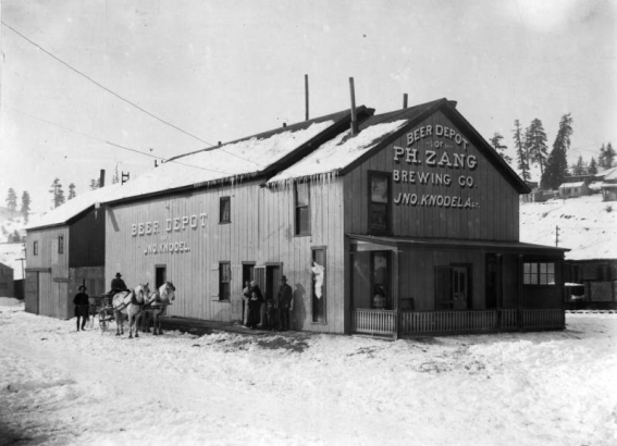 A wagon drawn by a team of horses is parked next to a brewery in the town of Creede, Colorado, in Mineral County. The driver sits in the cart and a man stands next to the wagon. The building is a two-story, wood- frame structure with gables, shingles, chimneys, rectangular windows, and a covered porch with spindles. Snow covers the ground and the hills in the background. A "Beer Depot of P.H. Zang Brewing Co. Jno. Knodel, Agt." shows.