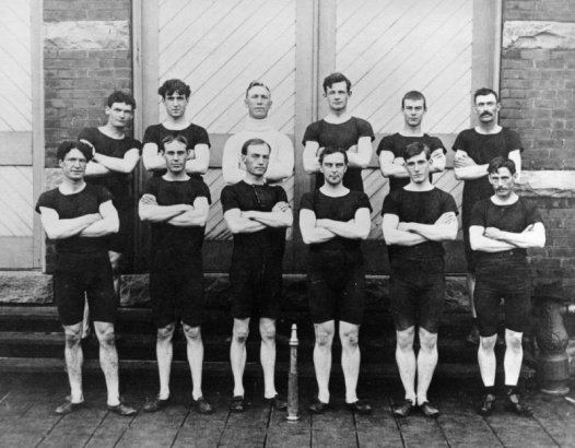 Colorado City Hose Team pose in front of the diagonal batten door outside the brick fire station, South Twenty-sixth and Cucharas streets, El Paso County, Colorado. The men stand wearing dark underwear shirts and shorts with arms crossed and their brass hose nozzle proudly on display in front of them.