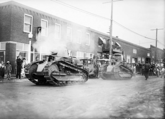 Two military tanks parade on a Arvada street, Jefferson County, Colorado. The lead tank's cannon smokes; drivers can be seen through small  front hatch doors. Arvada Chamber of Commerce sponsors the parade during the third Annual Harvest Festival. Spectators look on from the sidewalk and automobiles are parked curbside.