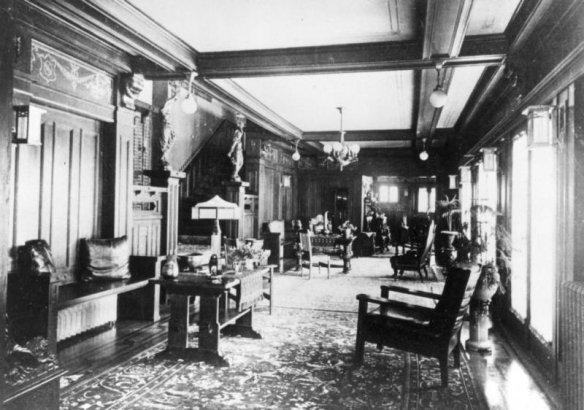 Interior view of the long reception hall in the Frederick Warshauer residence in Antonito, Conejos County, Colorado; includes Arts and Crafts furniture, chairs, tables, benches with leather pillows, lamps, hanging lanterns, oriental rugs, bronze sculptures, and potted palm plants; beamed ceiling with a stenciled border and stairway leading to the second floor.