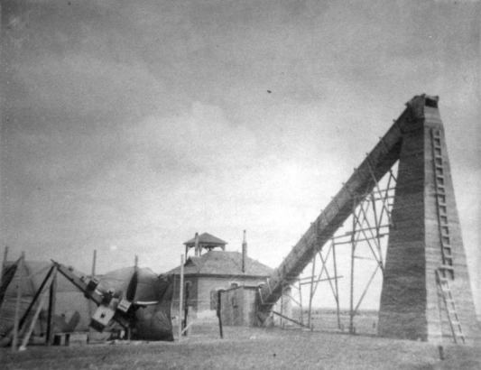 A telescope larger than the Brandon schoolhouse has a large tower with ladders attached to the side of it and a tube that forms an angle with the schoolhouse. Another mechanical device is attached to the one- story schoolhouse.