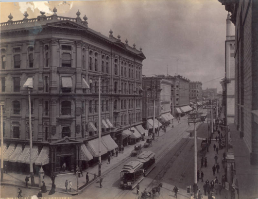 View of 16th (Sixteenth) and Larimer Streets in Denver, Colorado. Shows the Tabor Block and the Daniels and Fisher Dry Goods Store at 16th and Lawrence Streets. Signs read: "Rose & Co. Photographers," "Hotel Brunswick," and "United States Depository." A Denver Tramway Company trolley, carriages and pedestrians are in the street.