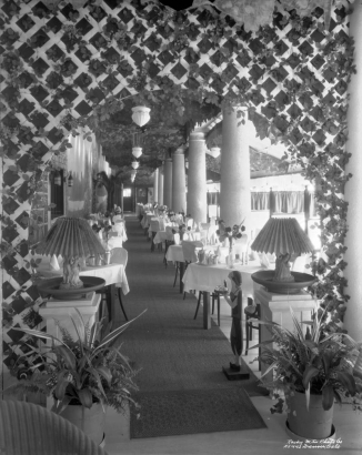 View of a restaurant at Lakeside Amusement Park in Lakeside (Jefferson County), Colorado; shows dining tables with tea service, flower vases, lattices, columns, ivy, potted plants, and ornate lamps. A wooden silhouette women in flapper costume holds an ashtray.