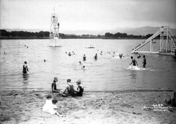Teenagers and girls play in the sand by Lake Rhoda at Lakeside Amusement Park in Lakeside (Jefferson County), Colorado; a lifeguard on a tower watches swimmers.