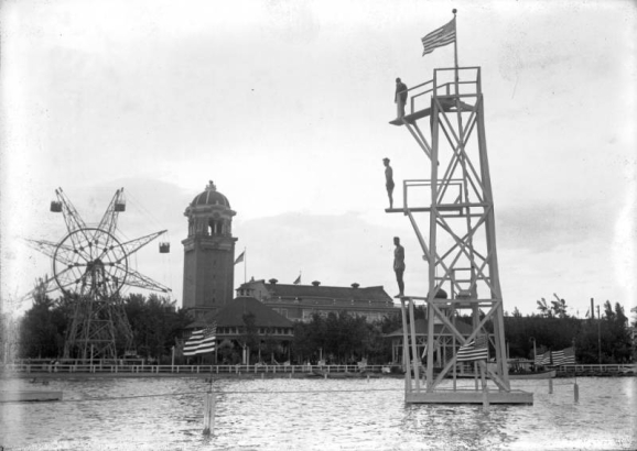 Men stand on a diving tower in Lake Rhoda at Lakeside Amusement Park in Lakeside (Jefferson County), Colorado; United States flags, the pavilion, and a ferris wheel are in the background.