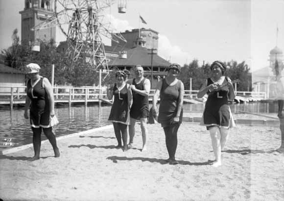 Women and a man walk in sand by Lake Rhoda at Lakeside Amusement Park in Lakeside (Jefferson County), Colorado; they wear swimming suits. One woman plays a ukelele.
