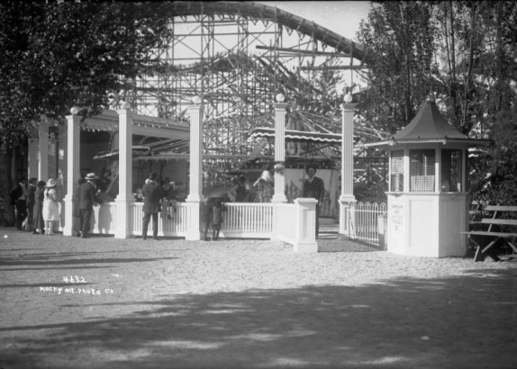 View of a ticket booth and roller coaster at Lakeside Amusement Park in Lakeside (Jefferson County), Colorado; people stand by railings.