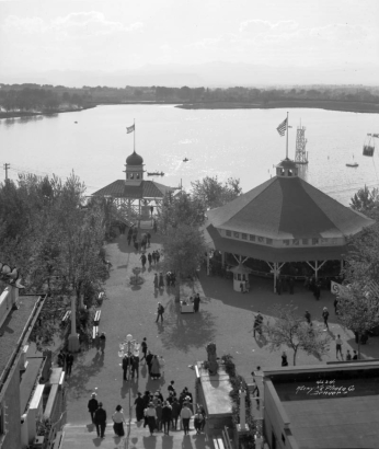 View of Lakeside Amusement Park in Lakeside (Jefferson County), Colorado; people walk by a pavilion and the boat house. Lake Rhoda and Lakewood are in the background.