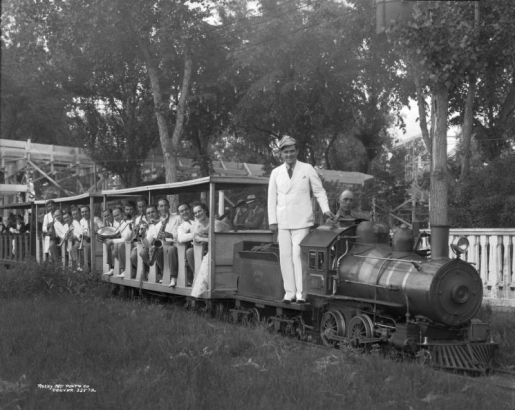 Jimmy Garrigan stands on a miniature locomotive at Lakeside Amusement Park in Lakeside (Jefferson County), Colorado; men and women with a guitar, saxophone, trombones and drums ride passenger cars.